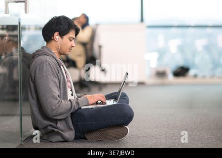Mixed-race male employee wearing earphones typing on laptop sitting with crossed legs on floor in office Stock Photo