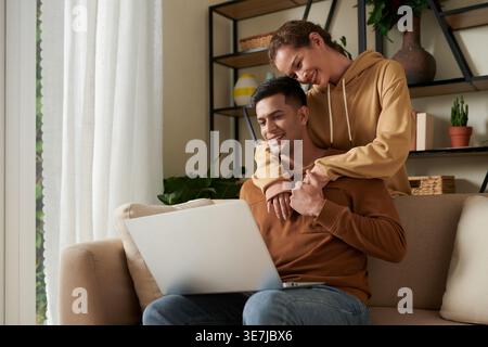 Smiling mixed race woman standing and embracing Asian boyfriend from behind looking at laptop sitting on sofa in living room at home Stock Photo