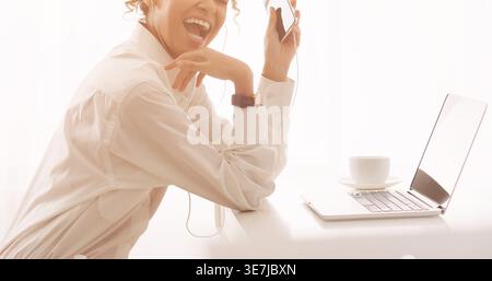 Cheerful Black business woman with curly hair listening to music sitting on front of laptop in office during daytime Stock Photo