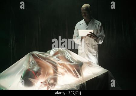 Black male scientist standing and using tablet examining caucasian woman lying with plastic cover on table in laboratory Stock Photo