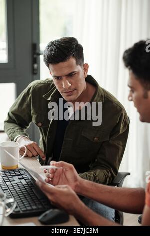 Indian male programmer discussing new program with male colleague while sitting at desk in office Stock Photo