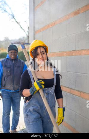 Hispanic female worker wearing denim overalls, yellow hardhat, and protective gloves walking carrying shovel on shoulder followed by Black foreman ... Stock Photo