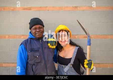Happy Hispanic female worker wearing denim overalls, yellow hardhat, and protective gloves holding shovel standing with arm on the shoulder of Blac... Stock Photo