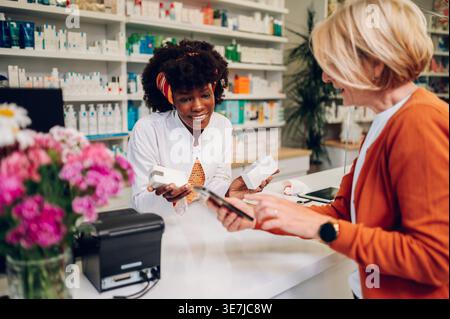 Black female pharmacist with afro holding 2 boxes standing at the counter against shelves with medical products talking to middle-aged caucasian fe... Stock Photo
