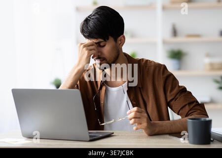 Tired indian man sitting in front of laptop at office, touching his head, suffering from burnout or financial crisis, young guy freelancer having diff Stock Photo