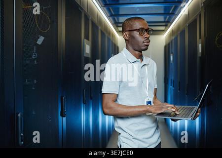 Black male IT engineer with eyeglasses looking at server cabinet holding laptop standing in data center Stock Photo