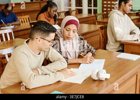 Middle Eastern male and Hispanic Muslim female immigrants studying together while sitting at desk in library Stock Photo