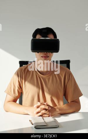 Asian man in orange t-shirt wearing VR headset sitting at the table at home Stock Photo
