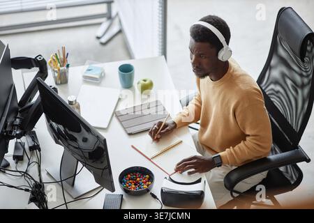 Black male programmer wearing headphones looking at computer monitor and writing on notebook while sitting at table in office Stock Photo