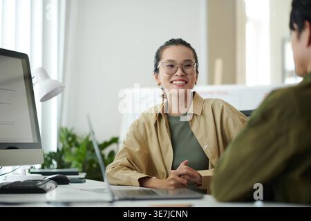 Asian female programmer with hands together wearing eyeglasses talking to male colleague sitting at table with laptop in office Stock Photo