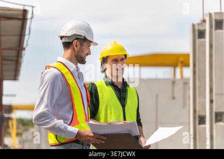 Hispanic male engineer with hardhat and safety vest discussing project with caucasian male colleague in construction site Stock Photo