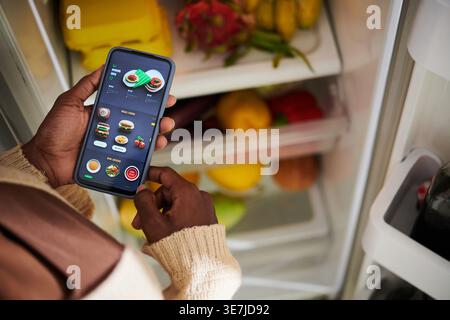 Black woman using smartphone with opened cooking application finding ingredients from fridge standing in kitchen at home Stock Photo