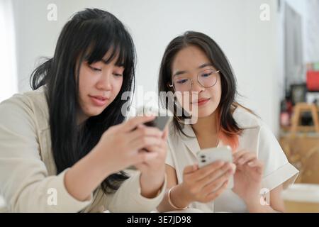 Asian female college students searching information on smartphones about homework sitting at table with cup of coffee and notebooks in cafe Stock Photo