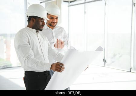 Diverse group of male engineers wearing hard hats standing together and looking at blueprint during work on construction site Stock Photo