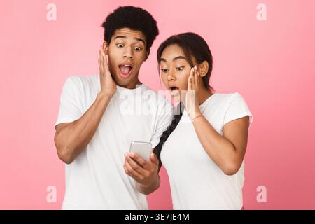 Shocked Black couple using smartphone while standing against pink background in studio Stock Photo