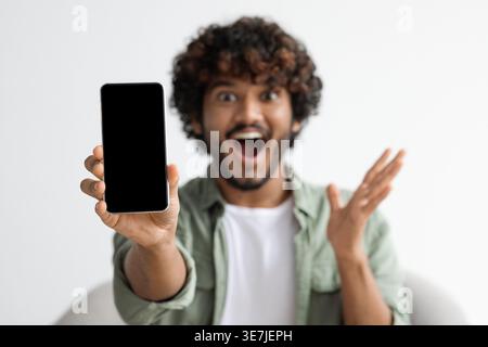 Shocked Asian man with curly hair holding and showing smartphone screen while sitting on chair at home Stock Photo