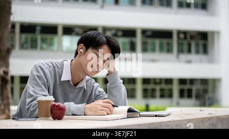 Asian male student leaning head on hand tired and sleepy sitting with books at table on campus during daytime Stock Photo