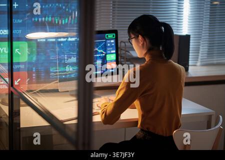 Focused Asian business woman wearing eyeglasses working on computer preparing for presentation sitting at table in office at night Stock Photo