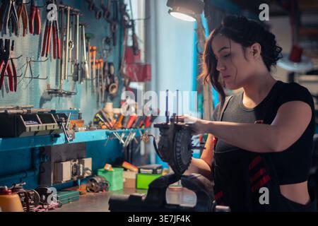 Hispanic female mechanic in messy bun wearing apron examining car parts standing in auto repair shop Stock Photo