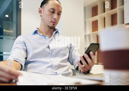 Frowning mixed-race businessman holding documents using smartphone sitting at table in office Stock Photo