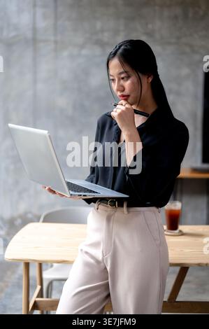 Thai business woman reading something on laptop screen standing in her modern office Stock Photo
