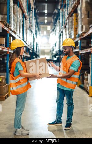 Black male worker wearing hard hat and safety vest scanning box held by caucasian female coworker while standing in warehouse Stock Photo