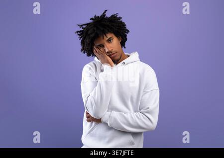 Black man with afro wearing white hoodie leaning head on his hand while standing against purple wall in studio Stock Photo