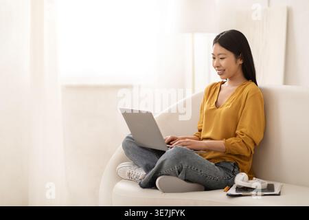 Positive smiling pretty young asian woman wearing casual outfit sitting on couch in living room, working from home, using laptop, chinese lady freelan Stock Photo