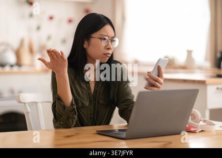 Irritated confused young asian lady wearing outfit and eyeglasses sitting at kitchen desk in front of laptop, working from home, looking at phone scre Stock Photo