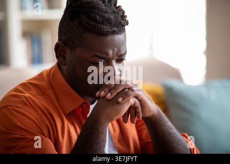 Depressed Black man leaning mouth on clasped hands sitting on sofa in living room at home Stock Photo