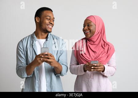 Black man and muslim wife in hijab holding smartphones and looking at each other while standing against gray background in studio Stock Photo