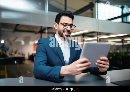 Middle Eastern man with beard and eyeglasses using tablet sitting at desk in office Stock Photo