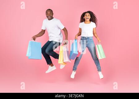 Cheerful Black couple with colorful shopping bags jumping together against pink background in studio Stock Photo
