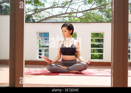 Asian woman meditating in lotus position and looking at tablet while sitting on yoga mat in balcony at home Stock Photo