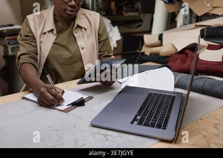 Black female tailor wearing eyeglasses holding tablet making notes while sitting at table with laptop in workshop Stock Photo
