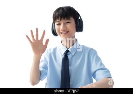 Smiling Chinese boy student in headphones waving hand while sitting at table against white background in studio Stock Photo