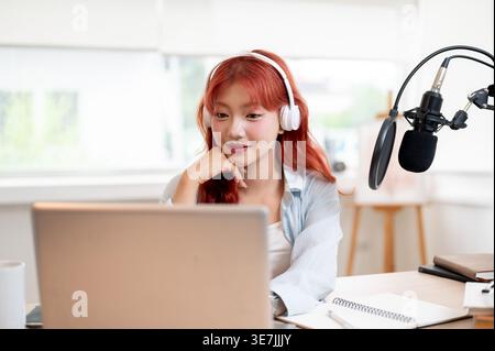 Asian female podcaster with headphones working on laptop while sitting at table with microphone in home studio Stock Photo