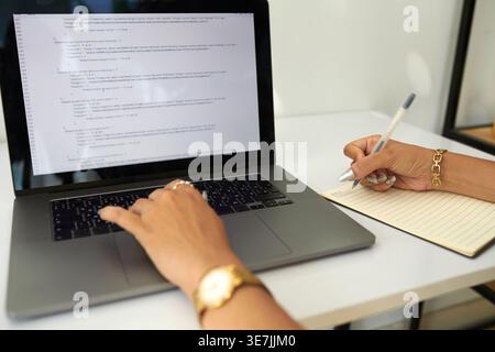 Hispanic woman using laptop and writing down notes while sitting at table in office Stock Photo
