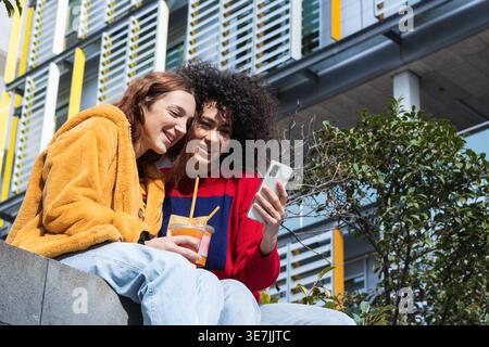 Hispanic woman with curly hair and caucasian female friend holding juice and using smartphone together while sitting on bench near building in city... Stock Photo