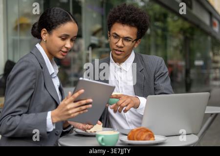 Black male and female business partners discussing project details holding cup of coffee and using tablet while sitting together at table with past... Stock Photo