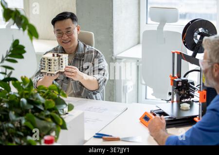 Asian male engineer checking building scale model while sitting at table with caucasian male colleague working on 3D printer in office Stock Photo