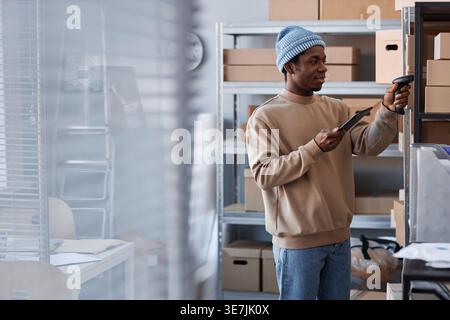 Black male worker wearing beanies checking package using tablet while standing near glass window in warehouse Stock Photo