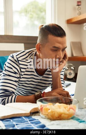 Vietnamese man leaning chin on hand working on laptop lying on bed with potato chips in bedroom at home Stock Photo