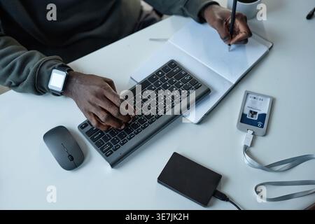 Hands of Black male programmer typing on computer using keyboard while taking notes sitting at table in office Stock Photo