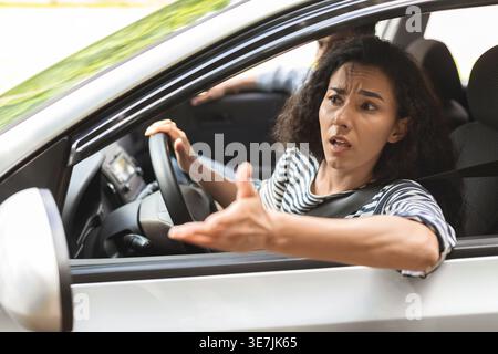 Middle Eastern woman with curly hair looking through window showing hand gesture while sitting inside car with passenger boyfriend during sunny day Stock Photo