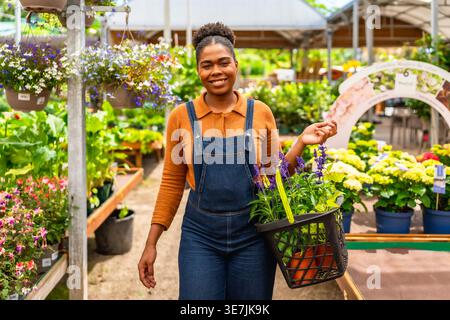 Hispanic female gardener in denim overalls holding basket with plants and smiling while standing in greenhouse Stock Photo