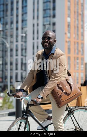 Black businessman wearing sunglasses riding bicycle on city street going to work on sunny day Stock Photo