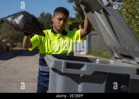 Hispanic male garbage collector throwing garbage bag in dumpster while standing in city street during sunny day Stock Photo