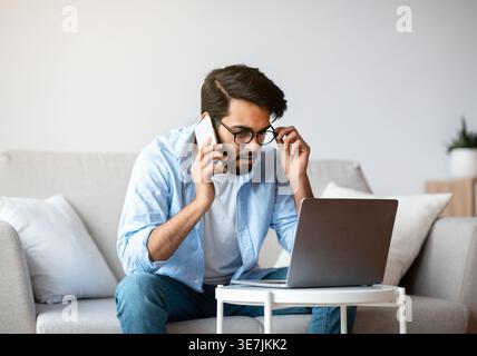 Arab Freelancer Man Talking On Cellphone And Working With Laptop At Home Office, Busy Eastern Guy Looking At Computer Screen While Discussing Project Stock Photo