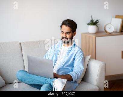 Eastern Millennial Male Using Laptop While Relaxing On Couch At Home. Handsome Arab Freelancer Guy Sitting On Sofa With Computer, Working Remotely Or Stock Photo